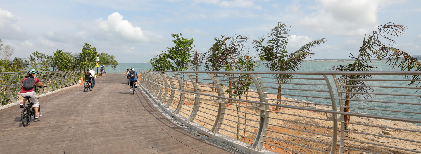 People cycling on wooden path next to metal railing, beach, and ocean under cloudy sky.